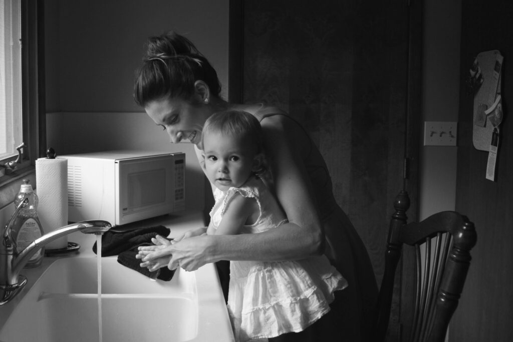 Black and white photo of a mother teaching her toddler to wash hands at a kitchen sink.