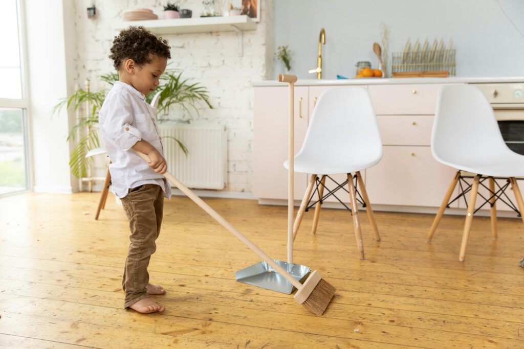 Young child sweeping wooden floor in bright modern living room with white chairs.