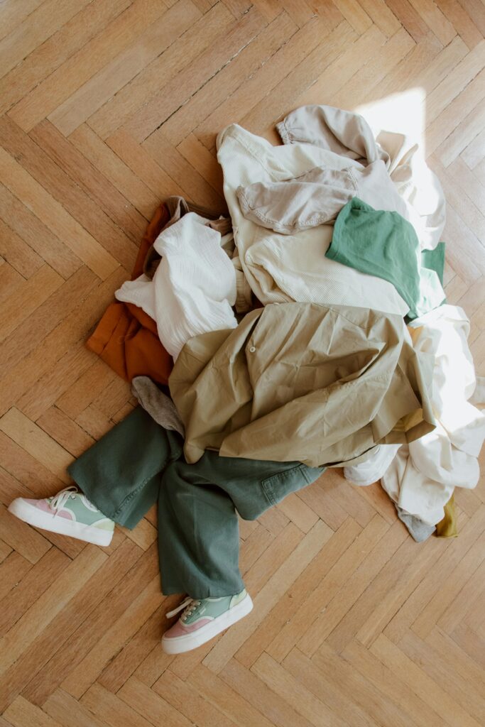 A child playfully hides under a pile of casual clothes on a parquet wooden floor.