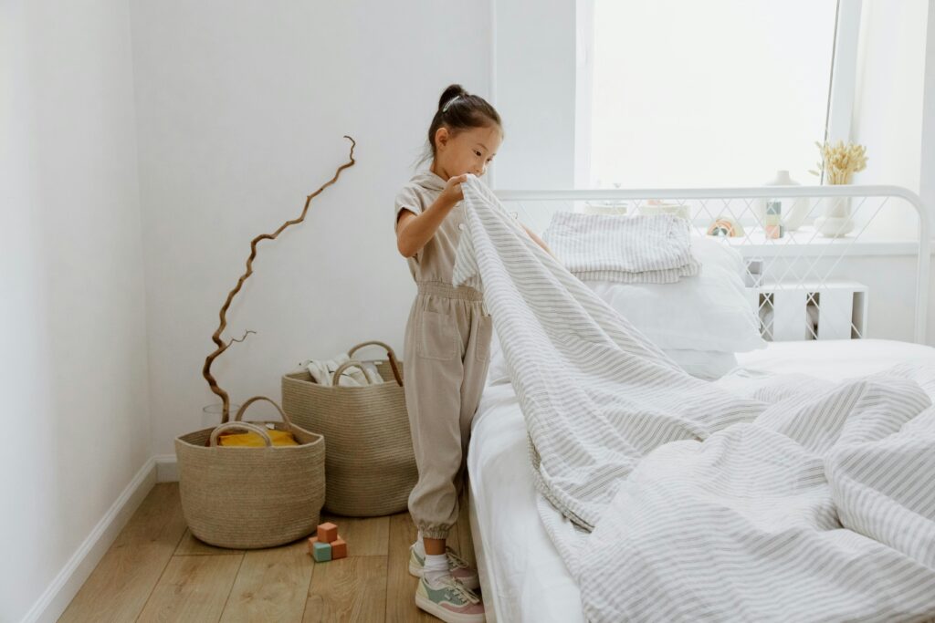 A young girl making her bed in a bright, minimalist bedroom.