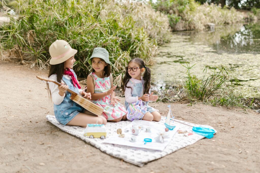 Three children enjoy a playful picnic by a pond, playing music and laughing together outdoors.