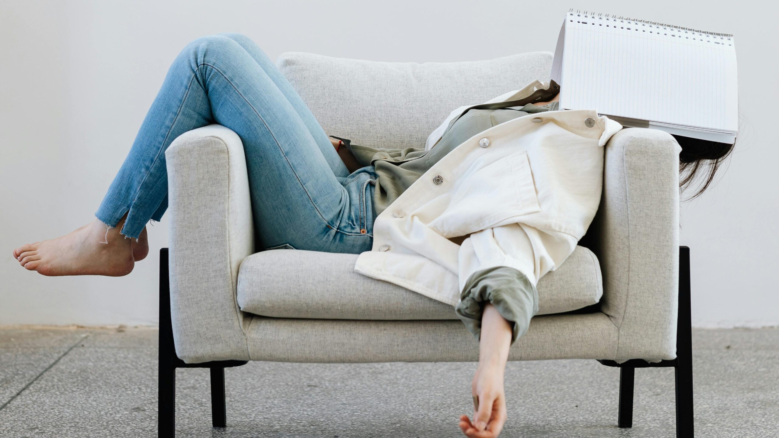 Tired woman in casual attire falls asleep on sofa with a notebook on face and pen in hand.